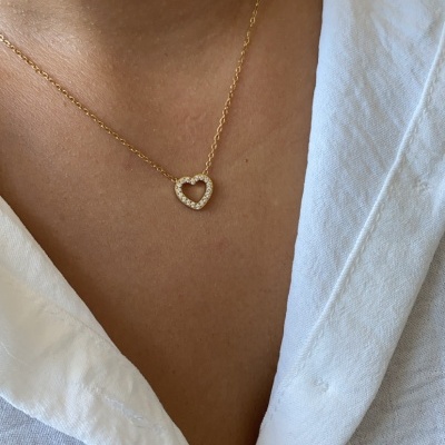 Colar dourado com pendente de coração com cristais sobre camisa branca.