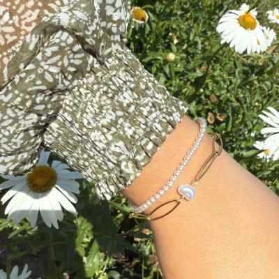 Pulseira com contas de pérolas e bracelete dourada com pingente de concha num pulso junto a flores margueritas