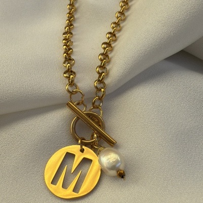 Colar dourado com pendente letra M e pérola sobre tecido creme.