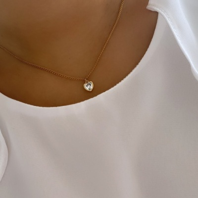 Colar dourado com pendente em forma de coração transparente numa pessoa com blusa branca