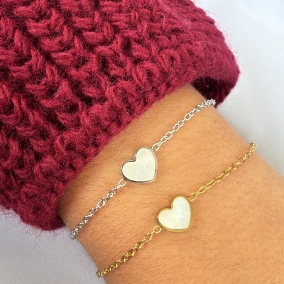 Duas pulseiras delicadas, prata e dourado, com corações brancos, no pulso de uma pessoa com camisola vermelha.