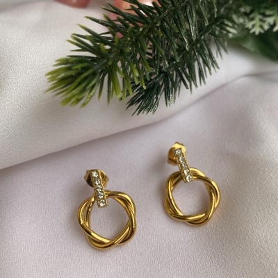 Brincos dourados entrelaçados com cristais sobre tecido branco e decoração verde e rosa