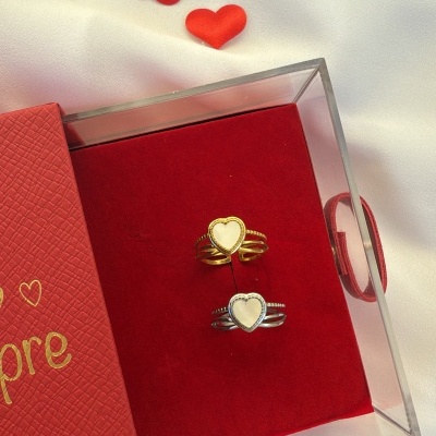 Dois anéis em forma de coração dentro de caixa transparente com base vermelha, caixa vermelha com a palavra Amore em dourado e corações vermelhos decorativos sobre tecido branco.