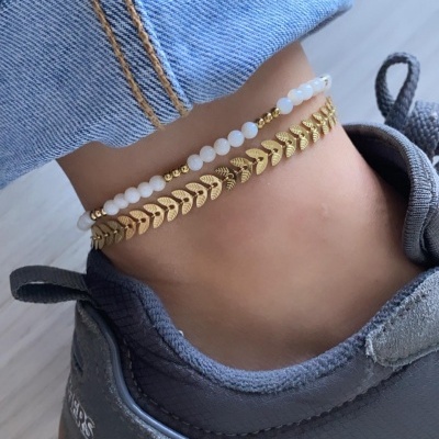 Tornozelo com duas pulseiras douradas e brancas, calçado desportivo cinzento e calças de ganga.