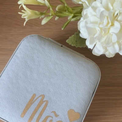 Estojo cinza claro com texto dourado 'Mãe' ao lado de flores brancas