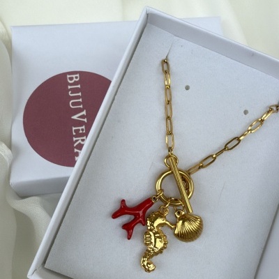 Colar dourado com pendentes de cavalo-marinho, concha e coral numa caixa branca com etiqueta BIJUVERA.
