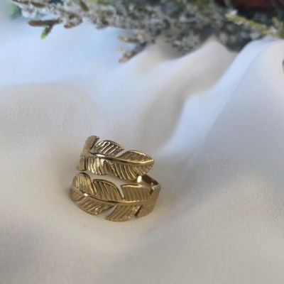 Anel dourado com design de folhas sobre tecido branco e ramos decorativos ao fundo