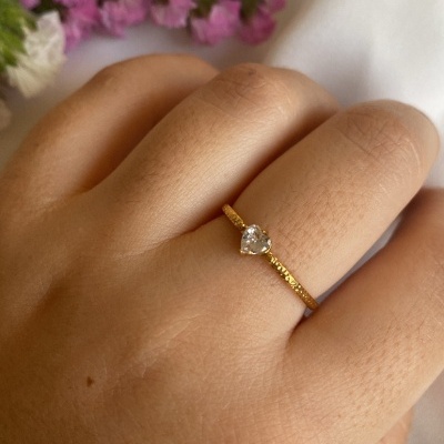 Anel dourado com pedra em forma de coração na mão, flores coloridas ao fundo