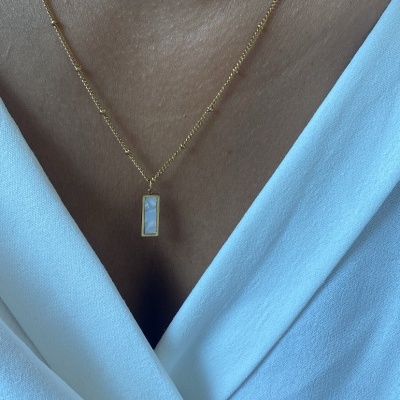 Colar dourado com pendente branco retangular num pescoço com camisa branca