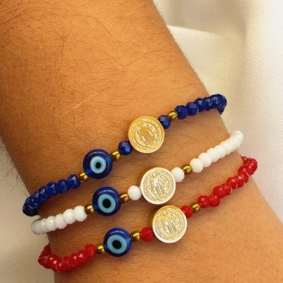 Três pulseiras coloridas com contas azuis, brancas e vermelhas, incluindo olhos gregos e moedas douradas, num pulso.