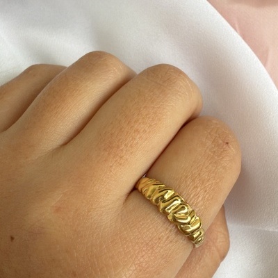 Anel dourado com textura irregular em dedo sobre tecido branco e rosa claro