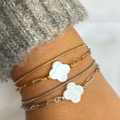 Pulseiras dourada e prateada com pendente de flor branca no pulso, com suéter cinza e fundo rosa e branco