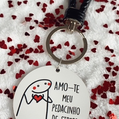 Chaveiro redondo branco com figura e texto, argola metálica e cordão preto em fundo com corações vermelhos