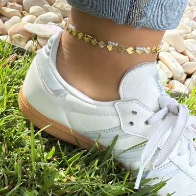 Ténis brancos em pele com sola castanha e tornozeleira dourada na perna sobre relva e pedras brancas
