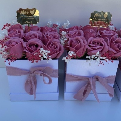 Dois arranjos florais em caixas brancas com laços cor-de-rosa e uma placa dourada com texto para mãe.