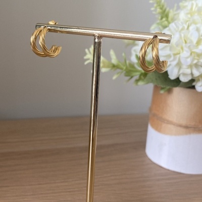 Brincos dourados em suporte metálico com base de madeira e planta ao fundo