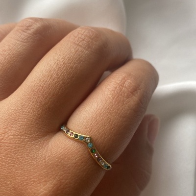 Anel dourado com pedras coloridas em formato de V no dedo médio, sobre tecido branco