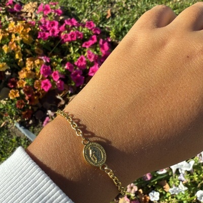 Pulseira dourada fina com pendente oval gravado num pulso com fundo de flores e relva