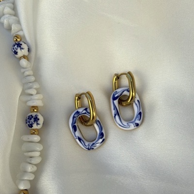 Brincos dourados com detalhes azul e branco e colar de contas brancas e douradas sobre tecido branco