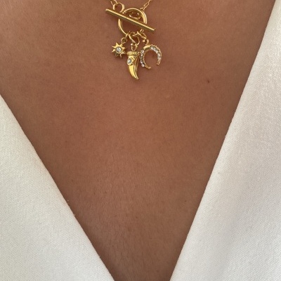 Colar dourado com pendentes de estrela, dente e ferradura no pescoço de pessoa de pele morena e roupa branca.