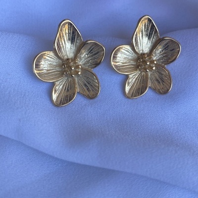 Brincos flor dourados sobre tecido branco com flores rosas ao fundo