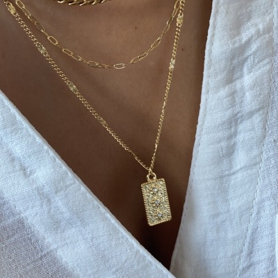Colar dourado em camadas com pendente retangular brilhante, usado sobre roupa branca com botão.
