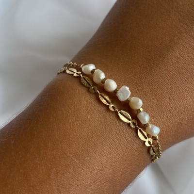 Bracelete duplo dourado com pérolas num pulso sobre fundo branco