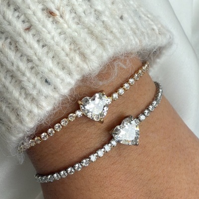 Pulso com duas pulseiras de brilhantes falsas com coração, uma dourada e outra prateada, e camisola branca de malha.