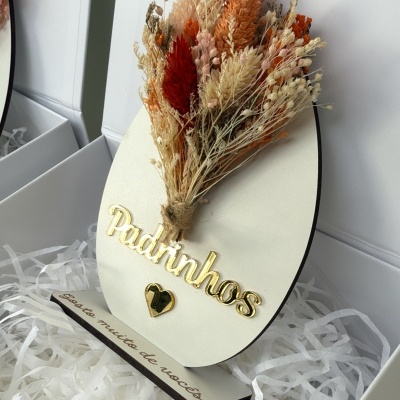 Objeto decorativo oval com flores secas e texto dourado Padrinhos numa caixa branca