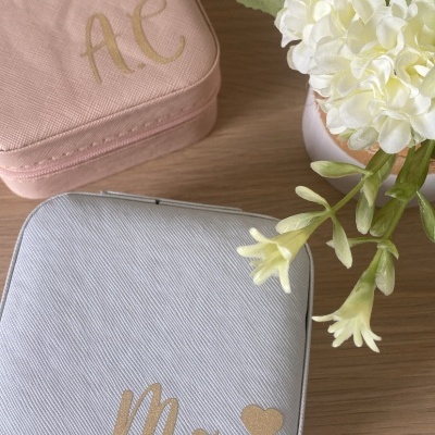 Duas bolsas quadradas pequenas, uma rosa com letras douradas AE e outra cinza com palavra Mãe e coração dourados, ao lado de flores brancas