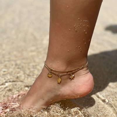 Tornozelo com pulseira dourada e pingentes na areia e água