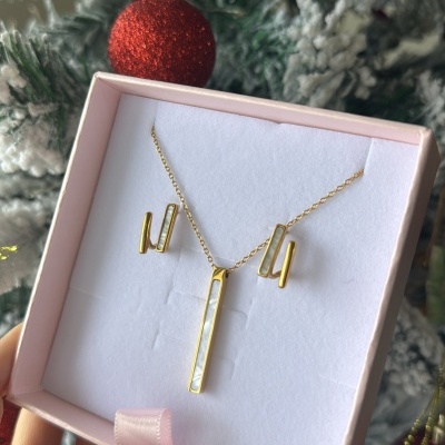 Conjunto de colar e brincos dourados com detalhes brancos numa caixa rosa, frente a árvore de Natal nevado com bolas vermelhas.