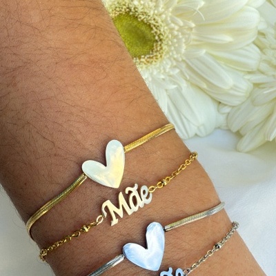 Dois conjuntos de pulseiras dourada e prateada com corações e palavra 'Mãe' e flor branca ao fundo