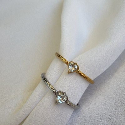 Dois anéis delicados com pedras em forma de coração, um dourado e outro prateado, sobre tecido branco.