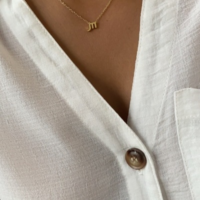 Colar dourado com letra m em corrente fina, camisa branca com botões castanhos.
