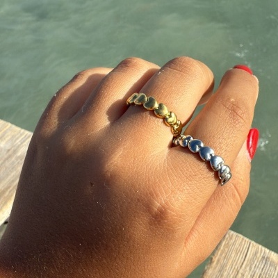 Mão com dois anéis de coração dourado e prateado e pulseira de contas rosas junto à água