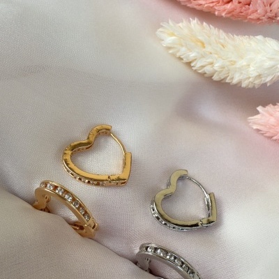 Brincos dourados e prateados de coração e com pedras sobre tecido acetinado creme com flores secas rosa e creme