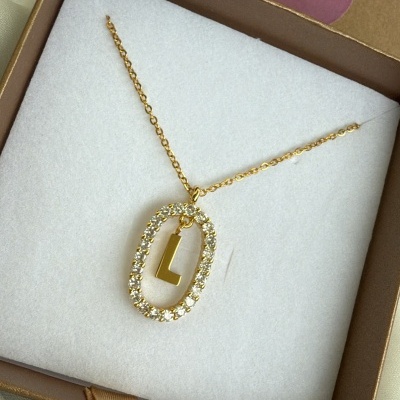 Colar dourado com pendente oval de pedras brilhantes e letra L numa caixa castanha com interior branco e etiqueta BIJUVERA