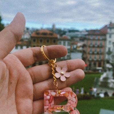 Chaveiro rosa em forma de letra B com flor e detalhes dourados sendo segurado por mão, fundo de jardim e prédios desfocados.