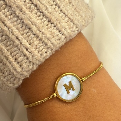 Pulseira dourada com medalha redonda e letra M ao centro num pulso com manga de malha bege.