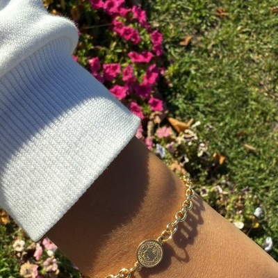 Pulseira dourada com pendente circular em pulso contra flores e relvado