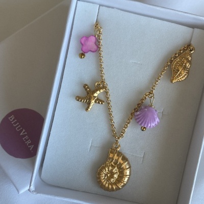 Colar dourado com pendentes de concha, estrela do mar e detalhes rosa numa caixa branca com etiqueta 'BIJURERA'.