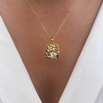 Colar dourado com pendente quadrado e pedra transparente sobre camisa branca