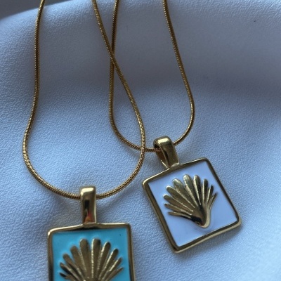 Dois colares dourados com pendentes quadrados azul claro e branco com conchas douradas em relevo sobre tecido branco