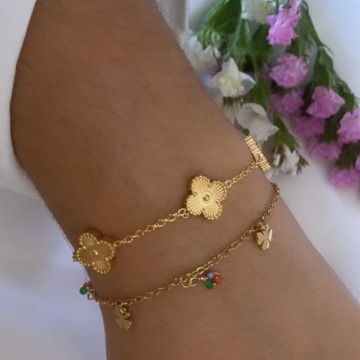 Pulseira dourada com flores e pendentes coloridos num pulso com camisa branca e flores ao fundo