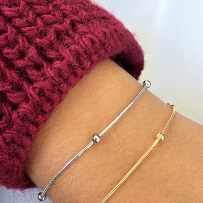 Pulseiras finas em prata e ouro com contas redondas num pulso. Roupas bordô e branco ao fundo.