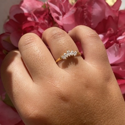 Anel dourado com cristais em forma de folhas num dedo com flores cor-de-rosa ao fundo