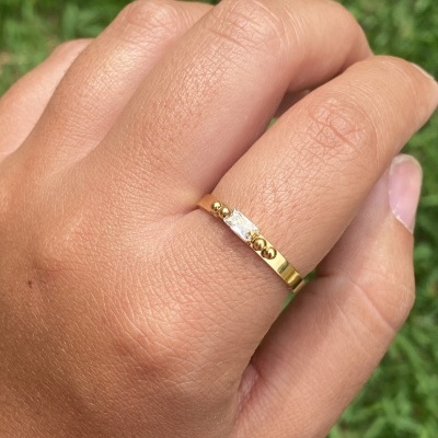 Anel dourado com pedra transparente e detalhes redondos numa mão sobre fundo de relva verde