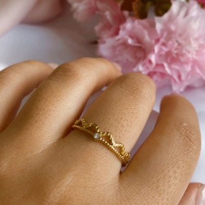 Anel dourado com design de coroa e pedra transparente num dedo com flores cor de rosa ao fundo