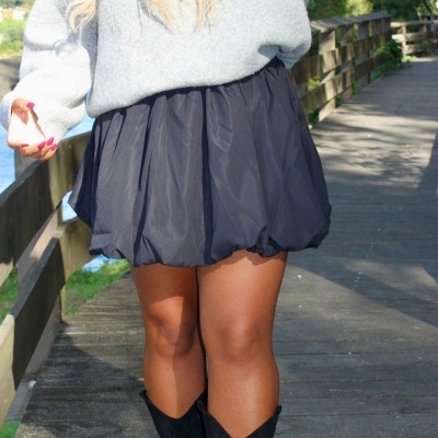 Mulher a usar saia preta curta, botas pretas altas e camisola cinzenta de malha num passadiço de madeira